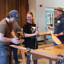 Load image into Gallery viewer, People carving paddles while laughing and smiling in a workshop.