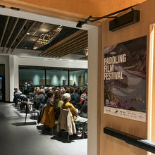 Load image into Gallery viewer, People watching a film in a room with a &#39;Paddling Film Festival&#39; poster on the door.
