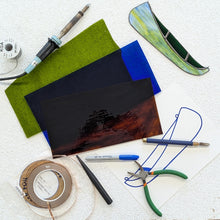 Load image into Gallery viewer, Workbench with stained glass tools and materials including a torch, pliers, and a piece of glass.
